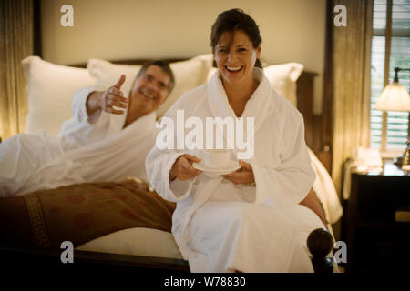 Porträt einer Mitte - erwachsene Frau sitzt auf einem Bett mit einer Tasse Kaffee zu, wie ihr Mann für sie in einem Hotel erreicht. Stockfoto