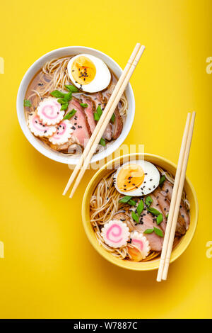 Zwei Schalen japanische Nudelsuppe "Ramen mit Fleischbrühe, geschnittenes Schweinefleisch, narutomaki, Eier mit Eigelb auf Pastell gelben Hintergrund. Traditionelles Gericht von Japan Stockfoto