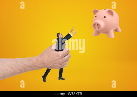 Big Hand kleine Unternehmer erreichen, mit seinen beiden Händen für niedliche rosa Sparschwein schwebend in der Luft auf gelben Hintergrund. Gebäude Karriere in f Stockfoto