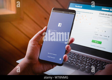 Bangkok, Thailand - August 5, 2019: Hände halten Smartphone mit Facebook auf dem Bildschirm und Facebook website auf Laptop Hintergrund anmelden. Facebook ist ein pop Stockfoto