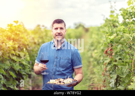 Gerne erfolgreiche Winzer im Weinberg holding Glas Rotwein und Board mit Käse, Nüssen und Trauben - lächelnde Menschen auf der Farm house Weingut Weinprobe Stockfoto