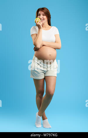 Leidenschaftlich gerne Fröhliche schwangere Frau Essen Vitamin, grüner Apfel. in voller Länge Foto,. Isolierte blauen Hintergrund. Studio erschossen. Gesundheit, Körperpflege. Stockfoto