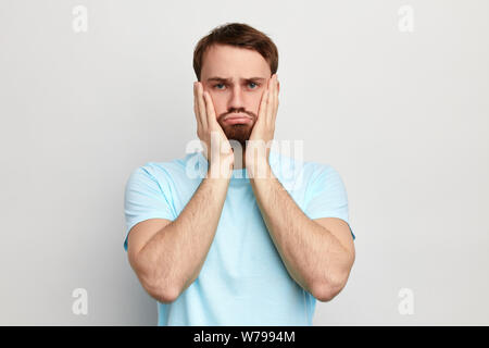 Deprimiert junger Mann mit blauen gemusterten männlichen halten Palmen auf Wange, Stirn sein Gesicht, mit traurigen Ausdruck an Kamera, Depression Konzept isoliert. Stockfoto