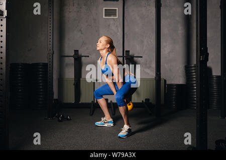 Sportlich motivierte starke Frau Trainieren mit einem Wasserkocher Bell, während er in Hocke. blondes Mädchen tun Kreuz fit Workout im Fitnessstudio. in voller Länge sid Stockfoto