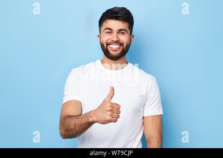 Happy bärtiger Mann im eleganten weißen T-Shirt mit strahlenden Lächeln zeigen, Daumen hoch, über blau Hintergrund isoliert, Studio erschossen. gute Arbeit, gut gemacht. vereinbart Stockfoto