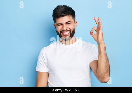 Gerne lustig, bärtiger Mann zwinkerte seinem Auge und zeigt ok Zeichen, auf blauem Hintergrund isoliert, alles unter Kontrolle ist. Witz, Spaß Konzept Stockfoto