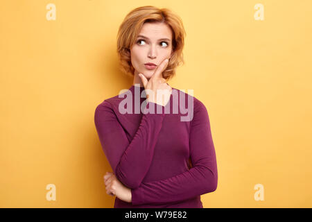 Stilvolle junge blonde Frau ihr Kinn berühren und seitlich mit zweifelhaft und skeptischen Ausdruck. Close up Portrait. gelb Zurück isoliert Stockfoto