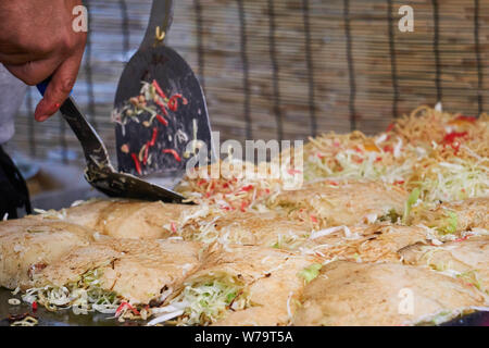 Die Hand des japanischen Mann benutzt Metall Spatel über Hiroshima - style Okonomiyaki um ein Essen bei einem Festival in Japan. Stockfoto