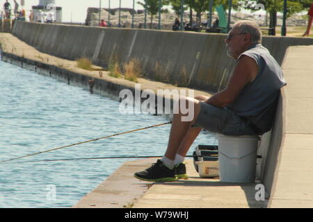 Ältere Mann auf einen Eimer Angeln am Lake Michigan in Kenosha, Wisconsin sitzen auf einem heißen Sommer morgen im August 2019. Stockfoto