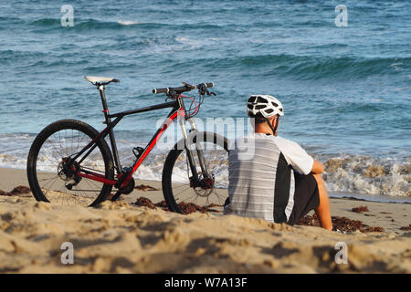 Ein Mann mit einem Fahrrad ist Ruhe am Strand des Atlantik. Sitzt im Sand. Ansicht von hinten. Stockfoto