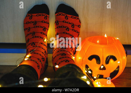 Kinder Beine in stilvollen warmen hellen bunt gestreiften lustige Socken in der girlande Lichter am Boden mit Kürbissen im Zimmer. Die Einrichtung für Halloween, gemütliche Moment. Gemütliche Stockfoto