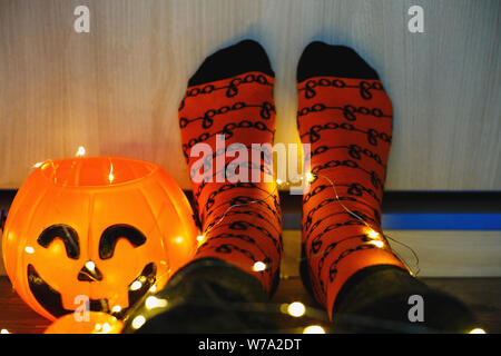 Kinder Beine in stilvollen warmen hellen bunt gestreiften lustige Socken in der girlande Lichter am Boden mit Kürbissen im Zimmer. Die Einrichtung für Halloween, gemütliche Moment. Stockfoto