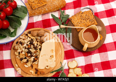 Picknick auf der Straße auf einem karierten Decke verteilt Essen. Honig und Nüssen auf einer hölzernen Fach, neben Brot, Käse, Tomaten und grünen. Stockfoto