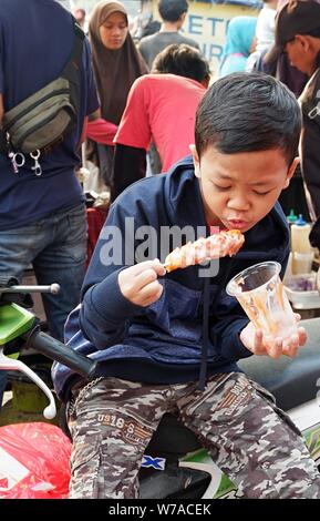 Jakarta, Indonesien - August 2019: ein Junge isst eine Wurst mit Stick vor einem Street Food stand. Stockfoto