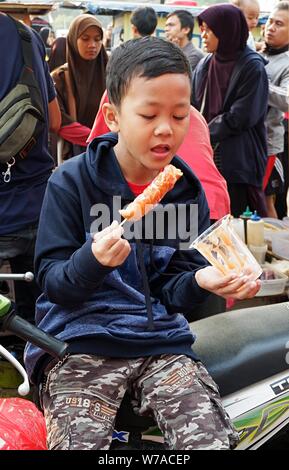 Jakarta, Indonesien - August 2019: ein Junge isst eine Wurst mit Stick vor einem Street Food stand. Stockfoto