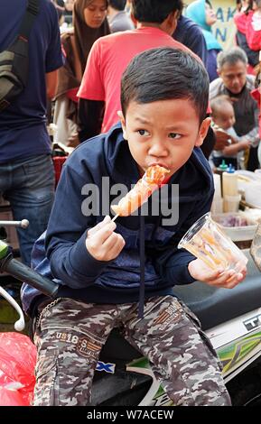 Jakarta, Indonesien - August 2019: ein Junge isst eine Wurst mit Stick vor einem Street Food stand. Stockfoto