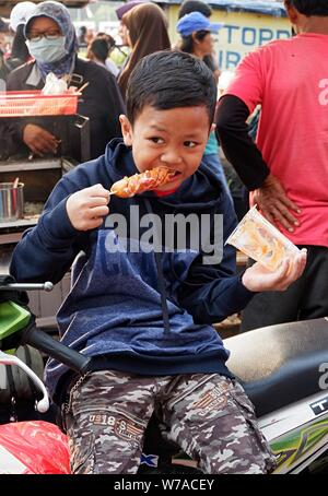 Jakarta, Indonesien - August 2019: ein Junge isst eine Wurst mit Stick vor einem Street Food stand. Stockfoto