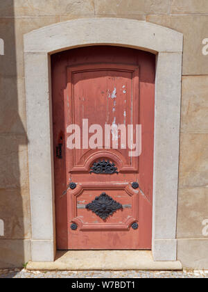 Eine Holztür mit verblasst und Peeling roter Farbe, mit Hoch dekorierte Metall Gartenmöbel set in eine Wand in den Gärten der Estoi Palace in Portu Stockfoto