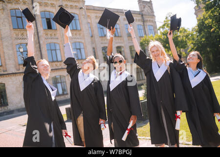 Fröhliche Absolventen erhöhen ihre Herren Caps in der Luft. Stockfoto