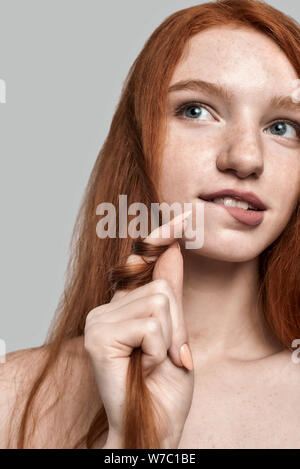 Studio geschossen von Jung und hübsch rothaarige Frau spielt mit ihren Haaren und Denken über etwas, während gegen grauen Hintergrund stehend Stockfoto