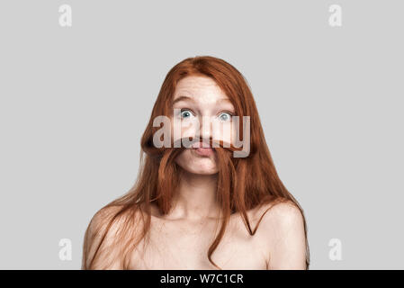 Studio shot Lustige junge rothaarige Frau spielt mit Ihrem Haar und einem Gesicht, während gegen grauen Hintergrund stehend Stockfoto