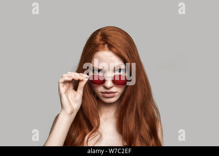 Schwere Dame. Portrait von selbstbewussten jungen rothaarigen Frau, die ihre Sonnenbrille einstellen beim Stehen gegen grauen Hintergrund. Studio shot Stockfoto