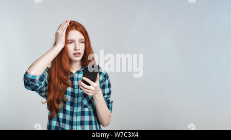 Studio geschossen von schockiert rothaarige Frau in Freizeitkleidung hand auf Kopf und Mobiltelefon gegen grauen Hintergrund stehen. Negative Emotionen. Digitale Stockfoto