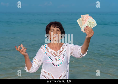 Eine ältere Frau hält ein Bündel von thailändischen Banknoten in die Hände und lächelte. Gerne ältere Frau mit viele Banknoten. Rentenfonds und Reisen Stockfoto
