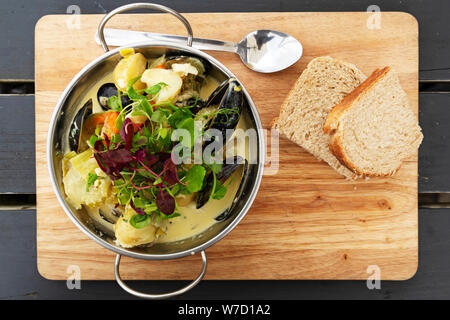 Eine Schüssel mit Seafood Chowder ist auf einem Holzbrett mit Brot serviert. Der Teller mit Kartoffeln und Salat. Stockfoto