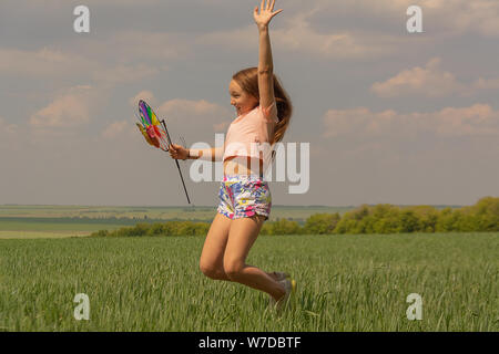 Glückliches Mädchen mit langem Haar mit einem farbigen Mühle Spielzeug in die Hände erhebt ihre Hand und springt. Konzept der Sommer, Freiheit und glückliche Kindheit. Stockfoto