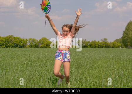 Glückliches Mädchen mit langem Haar mit einem farbigen Mühle Spielzeug in die Hände erhebt ihre Hand und springt. Konzept der Sommer, Freiheit und glückliche Kindheit. Stockfoto