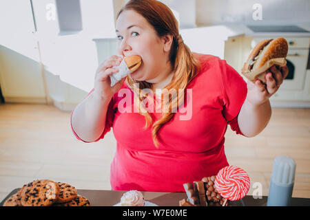 Fett junge Frau in der Küche sitzt und isst Junk Food. Ungesunde Lebensweise. Beißen Stück Burger und Blick auf Kamera. Körper positiv. Bonbons auf Tabl Stockfoto