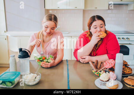 Junge hungrige Schlank und übergewichtige Frauen in der Küche essen. Gesundes und ungesundes Essen. Salat vs Burger. Körper positiv. Gesunder Lebensstil Stockfoto