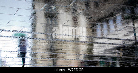 Unscharfe Reflexion Schatten Silhouette einer Frau alleine unter dem Dach an einem regnerischen Fußgängerzone Stadt nasse Straße an einem Sommertag, in einer Pfütze Stockfoto