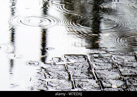 Detail der Regen fällt in eine Pfütze und Reflexionen über eine gepflasterte Straße Stockfoto