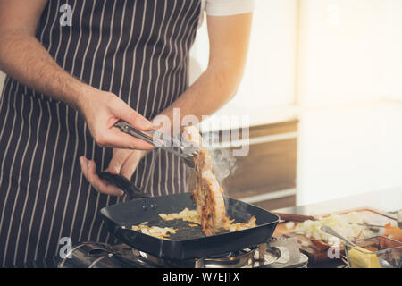 Nahaufnahme der Mann, der gestreifte Schürzen schelle Fleisch aus der Pfanne auf dem Gasherd in der Küche Stockfoto