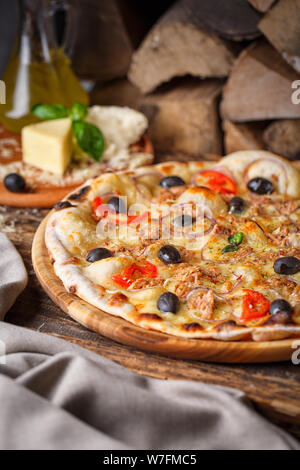 Pizza mit Fisch, Oliven, Zwiebeln und Tomaten auf dem Hintergrund des Brennholzes Stockfoto