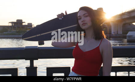 Junge attraktive Frau stehend auf der Uferpromenade am sunset Holding ein Skateboard - Posing auf der Kamera. Mitte der Schuß Stockfoto
