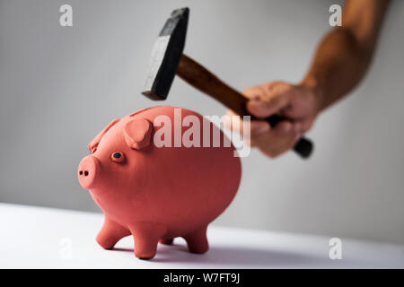 Nahaufnahme von einem kaukasischen Mann über zu brechen, indem sie mit einem Hammer, ein Sparschwein auf eine weiße Unterlage gebracht Stockfoto