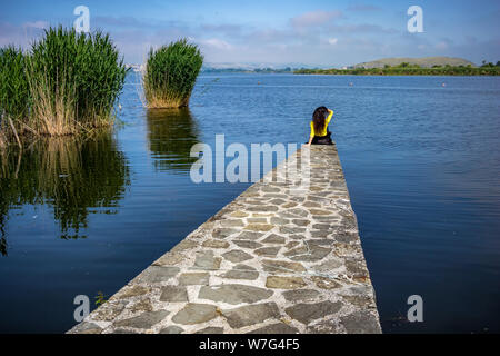 Dorf Seeblick von Ioannina, Griechenland mit unkenntlich Mädchen mit langen Haaren und gelbe Bluse sitzt auf Pier am See Pamvotida. Early morn Stockfoto