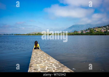 Dorf Seeblick von Ioannina, Griechenland mit unkenntlich Mädchen mit langen Haaren und gelbe Bluse sitzt auf Pier am See Pamvotida. Early morn Stockfoto