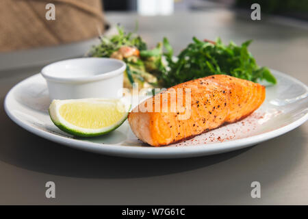 Gegrilltes Lachssteak auf der Seite mit Rucola und Salat. Mit einer Soße und Kalk auf eine weisse Platte im Restaurant serviert. Nahaufnahme. Stockfoto