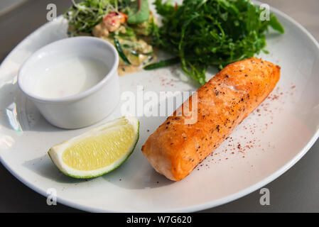 Gegrilltes Lachssteak auf der Seite mit Rucola und Salat. Mit einer Soße und Kalk auf einem weißen Teller serviert. Weiße Sauce. Stockfoto