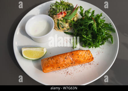 Gegrilltes Lachssteak auf der Seite mit Rucola und Salat. Mit einer Soße und Kalk auf einem weißen Teller serviert. Weiße Sauce. Stockfoto