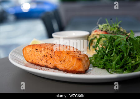 Gegrilltes Lachssteak auf der Seite mit Rucola und Salat. Mit einer Sauce auf einem weißen Teller im Restaurant serviert. Nahaufnahme mit Bokeh. Stockfoto
