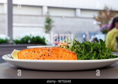 Gegrilltes Lachssteak auf der Seite mit Rucola und Salat. Mit einer Soße und Kalk auf eine weisse Platte im Restaurant serviert. Stockfoto