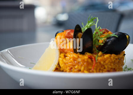 Spanische Paella mit Muscheln und Garnelen mit Zitrone und Rucola. Auf einem weißen Teller serviert. Nahaufnahme mit Bokeh. Main Reisgericht. Restaurant. Stockfoto