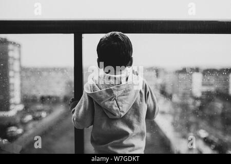 Schwarz-Weiß-Aufnahme eines Kindes, das ein graues trägt kapuzenpullover auf einem Balkon Stockfoto