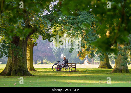 Northampton GROSSBRITANNIEN. 2. August 2019. Einen warmen, sonnigen Morgen für diese Radfahrer, der eine Entlüftung auf der Parkbank in Abington Park, lange Schatten caus Stockfoto