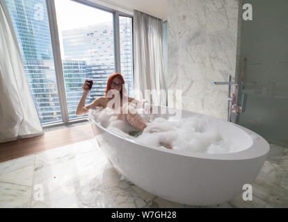 Eine rothaarige Frau im Schaumbad liegen und ein Glas Rotwein halten - Ein Blick auf die modernen Gebäude aus dem Panoramafenster Stockfoto